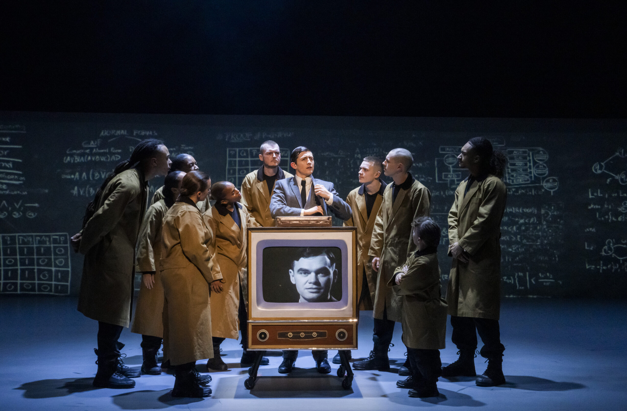 Photo by Tristram Kenton of a scene in Free Your Mind. Several individuals wearing brown trench coats look to a person in a suit and tie speaking. In front of the suit and tie person is a old style wood cabinet black and white television with a close of a man's face