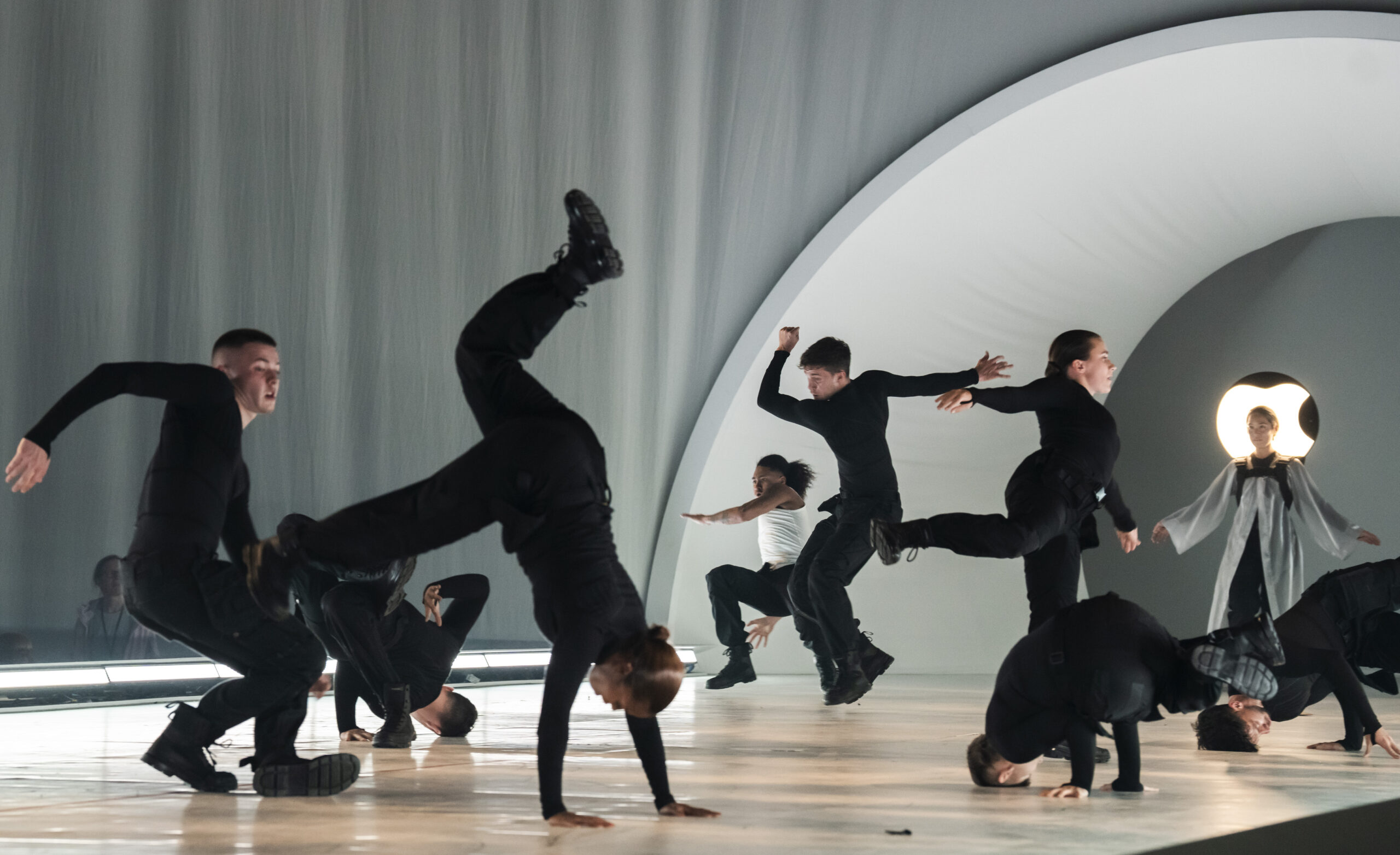 Photo by Photo by Tristram Kenton of Free Your Mind. Seven people in black outfits, one in a white sleeveless shirt, and one more in a white dress or robe dance on a wooden stage in front of gray curtains and a glowing white logo. Some are in mid-flip and mid-headspins