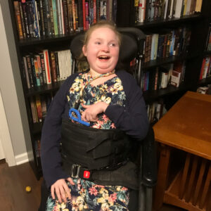 photo of Caitlin Thomas wearing a flower-print dress, sitting in her chair, and smiling. Shelves of books are behind her